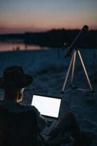 A person works on a laptop next to a telescope by a lake at twilight, blending nature and technology.